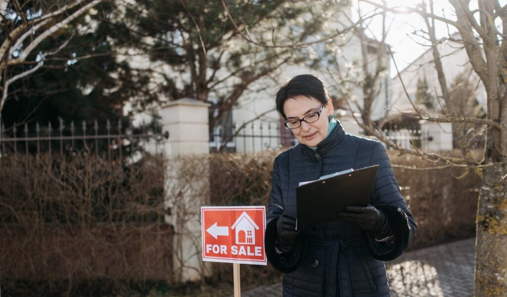 A Woman Standing Beside the House for Sale Signage